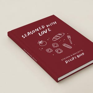 May include: A deep red hardcover recipe book with white text that reads Seasoned with Love and Steven & Bailey's Recipe Book. The cover features white line art illustrations of a loaf of bread, a tomato, a fork, and pasta shapes.