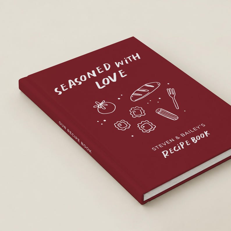 May include: A deep red hardcover recipe book with white text that reads Seasoned with Love and Steven & Bailey's Recipe Book. The cover features white line art illustrations of a loaf of bread, a tomato, a fork, and pasta shapes.