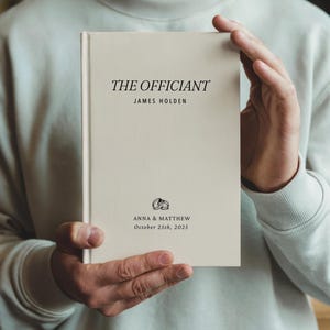May include: A cream-colored book titled "THE OFFICIANT" with the name "JAMES HOLDEN" printed in black. The bottom of the book features the names "ANNA & MATTHEW" and the date "October 25th, 2025", along with a graphic of two wedding rings.