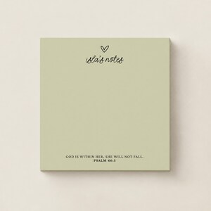 May include: A square sage green notepad featuring a small black heart icon above the text "isla's notes" in a cursive script. At the bottom, the text "GOD IS WITHIN HER, SHE WILL NOT FALL. PSALM 46:5" is printed in a clean black serif font.