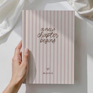 May include: A hardback journal with a striped design in pale pink and white. The cover displays the text "a new chapter begins" in a handwritten style, a heart symbol, and the name "Maisie".