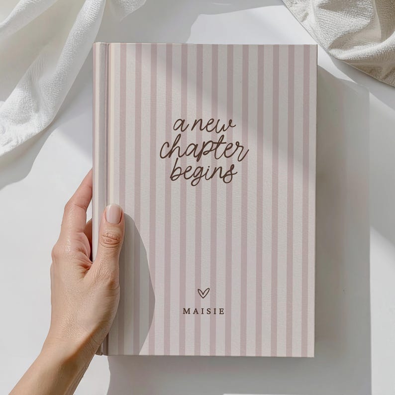 May include: A hardback journal with a striped design in pale pink and white. The cover displays the text "a new chapter begins" in a handwritten style, a heart symbol, and the name "Maisie".