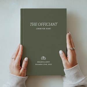 May include: An olive green book with the title "THE OFFICIANT" in white script. The name "JENNIFER HART" is below the title. The bottom of the book has the names "HELEN & ANDY" and the date "November 23rd, 2025".