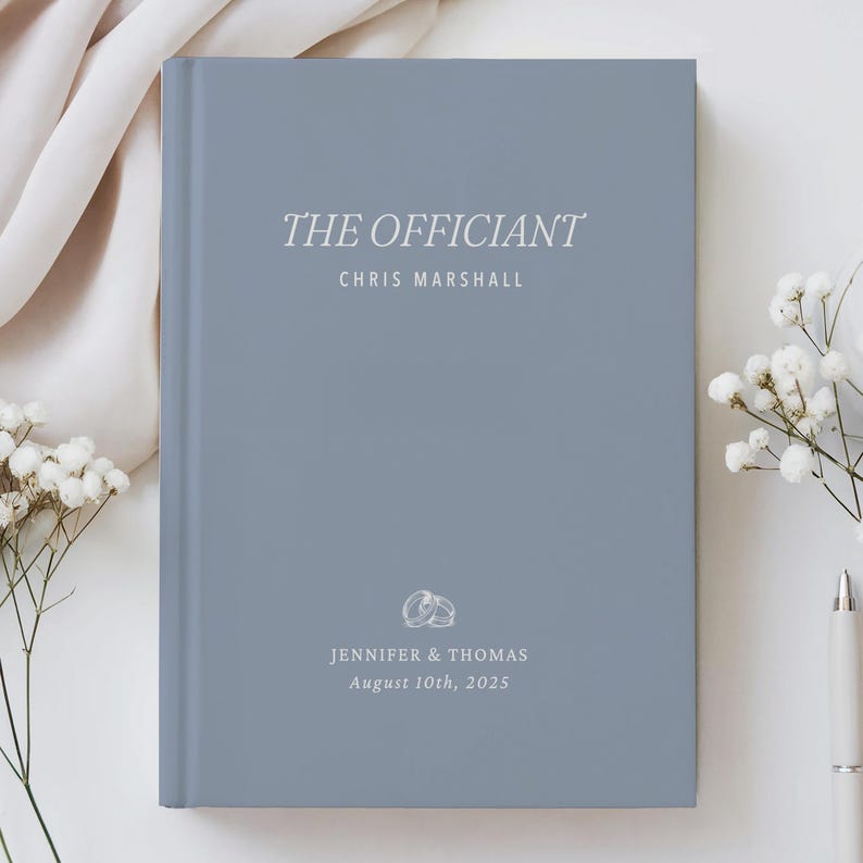 May include: A blue-gray book with the words "THE OFFICIANT" in elegant white script. Below, the name "CHRIS MARSHALL" is printed. The bottom of the book features the names "JENNIFER & THOMAS" and the date "August 10th, 2025", along with a ring graphic.