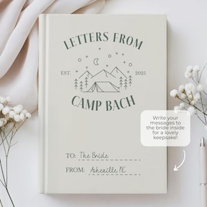 Puede incluir: Un libro blanco con un logotipo verde que dice "Letters From Camp Bach Est. 2025". El libro tiene una l&iacute;nea para "To: The Bride" y "From: Asheville, NC". El libro est&aacute; sobre una superficie blanca con flores blancas.