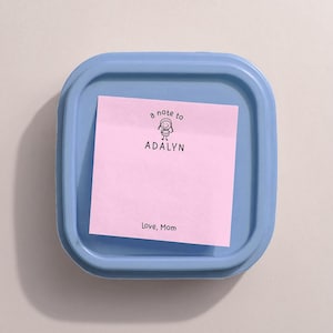 May include: A light blue square container with a pink sticky note inside. The note has the text "a note to ADALYN" with a small illustration of a person and the words "Love, Mom".