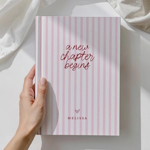 May include: A pink and white striped journal with the words "a new chapter begins" in burgundy script. A small heart and the name "Melissa" are below the text. The journal is held by a hand.