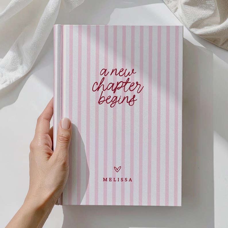 May include: A pink and white striped journal with the words "a new chapter begins" in burgundy script. A small heart and the name "Melissa" are below the text. The journal is held by a hand.