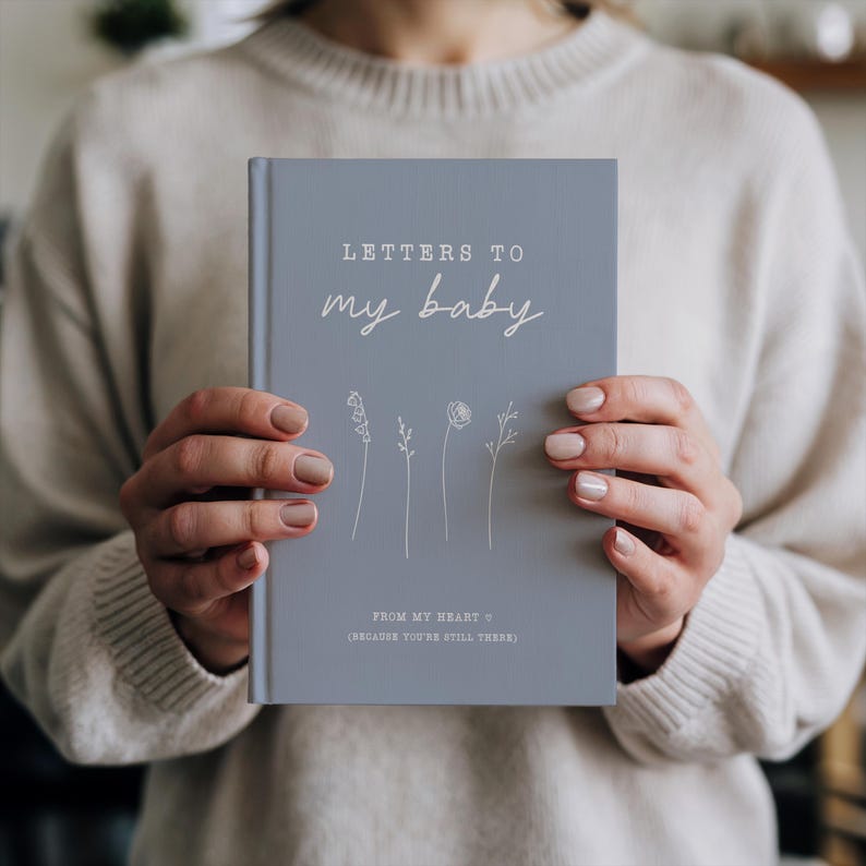 Op de afbeelding: Een blauw notitieboek met de tekst "Letters to my baby" en "From my heart (because you're still there)" in witte letters. Het notitieboek heeft een eenvoudig bloemenmotief.