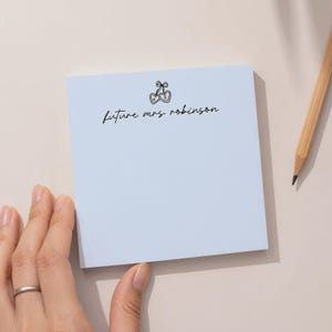 May include: A square light blue sticky note featuring a black line drawing of two connected hearts with a bow on top, containing the letters H and R. The text "future mrs robinson" is printed in black cursive script below the heart illustration.