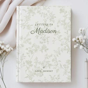 May include: A hardcover journal with a floral pattern in shades of green and white. The cover reads "Letters to Madison" in elegant script, with "Love, Mommy" below. The journal is surrounded by white flowers and a pen.