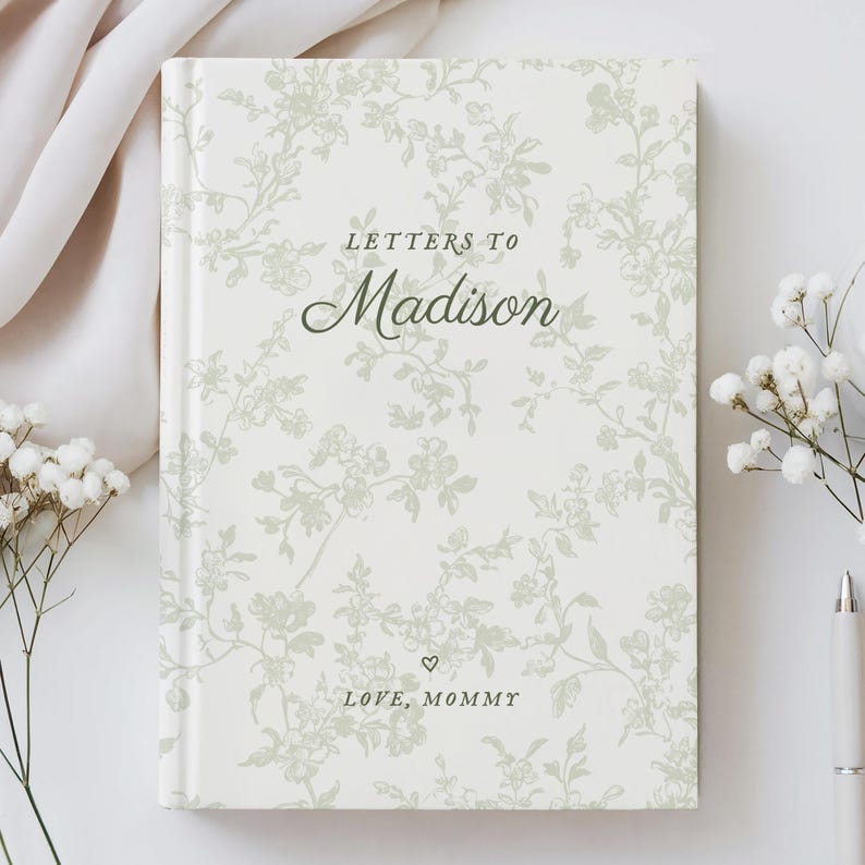 May include: A hardcover journal with a floral pattern in shades of green and white. The cover reads "Letters to Madison" in elegant script, with "Love, Mommy" below. The journal is surrounded by white flowers and a pen.