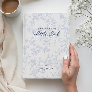 May include: A hardcover journal titled "Letters to My Little Girl" with a floral pattern in light blue and white. The cover includes the text "LOVE, MAMA" and a heart symbol. A white mug with coffee is in the upper left corner.