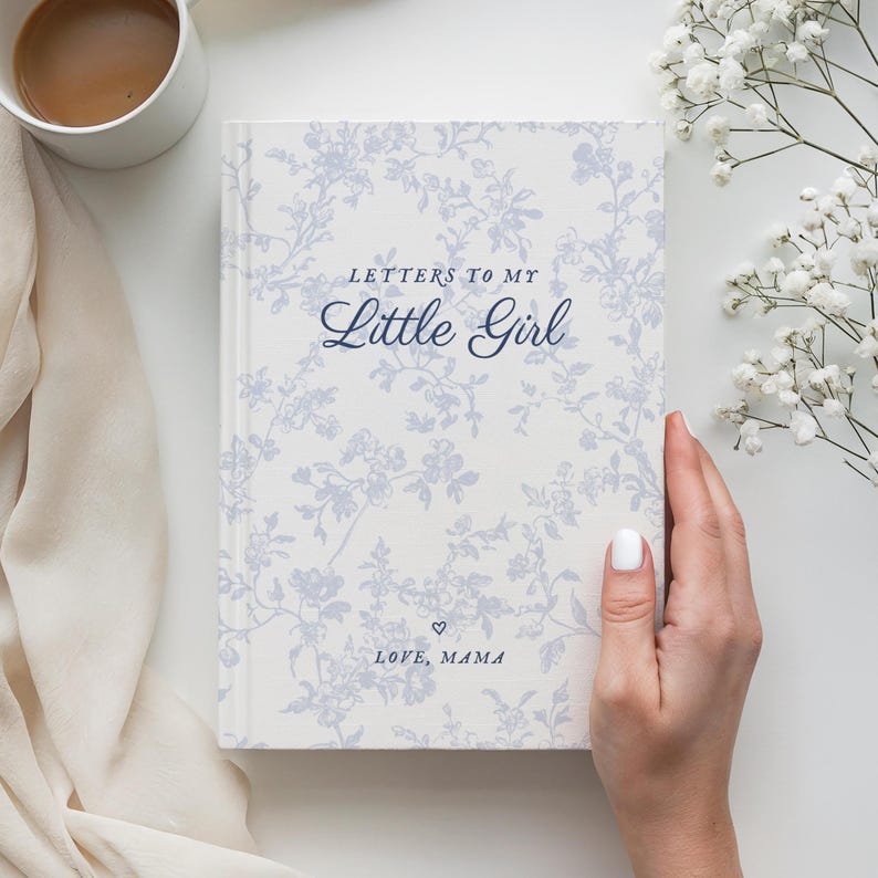 May include: A hardcover journal titled "Letters to My Little Girl" with a floral pattern in light blue and white. The cover includes the text "LOVE, MAMA" and a heart symbol. A white mug with coffee is in the upper left corner.