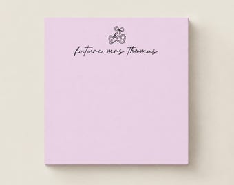 May include: A square light purple notepad featuring a black line drawing of two hearts tied with a bow at the top. Below the illustration, the text reads future mrs Thomas in a black cursive script font centered on the paper.