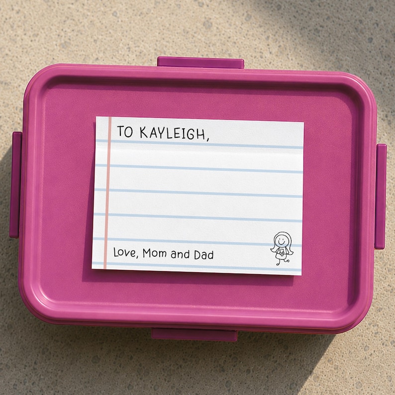 K&ouml;nnte beinhalten: Eine rechteckige magenta-farbene Brotdose aus Kunststoff mit einem wei&szlig;en Zettel auf dem Deckel. Der Zettel hat blaue horizontale Linien, einen rosa vertikalen Rand und den aufgedruckten Text: TO KAYLEIGH, Love, Mom and Dad, sowie eine kleine Strichm&auml;nnchen-Zeichnung.