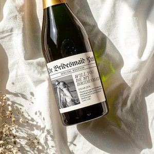May include: A champagne bottle with a custom label that reads "The Bridesmaid Post" and "Will You Be My Bridesmaid". The label includes a black and white photo and a personal message. The bottle has a gold foil top.