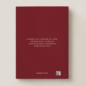 May include: A deep red rectangular book cover featuring centered white text that reads: CHEERS TO A LIFETIME OF LOVE, SERVED WITH A SIDE OF LAUGHTER AND A GENEROUS PORTION OF JOY! The bottom includes the text THOUGHT OF YOU and a small QR code.