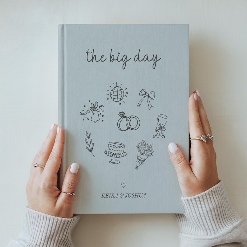Pu&ograve; includere: Un diario di nozze azzurro chiaro intitolato "the big day" con illustrazioni disegnate a mano a tema matrimonio. Il diario &egrave; tenuto in mano con anelli. I nomi "Keira & Joshua" sono stampati in basso.