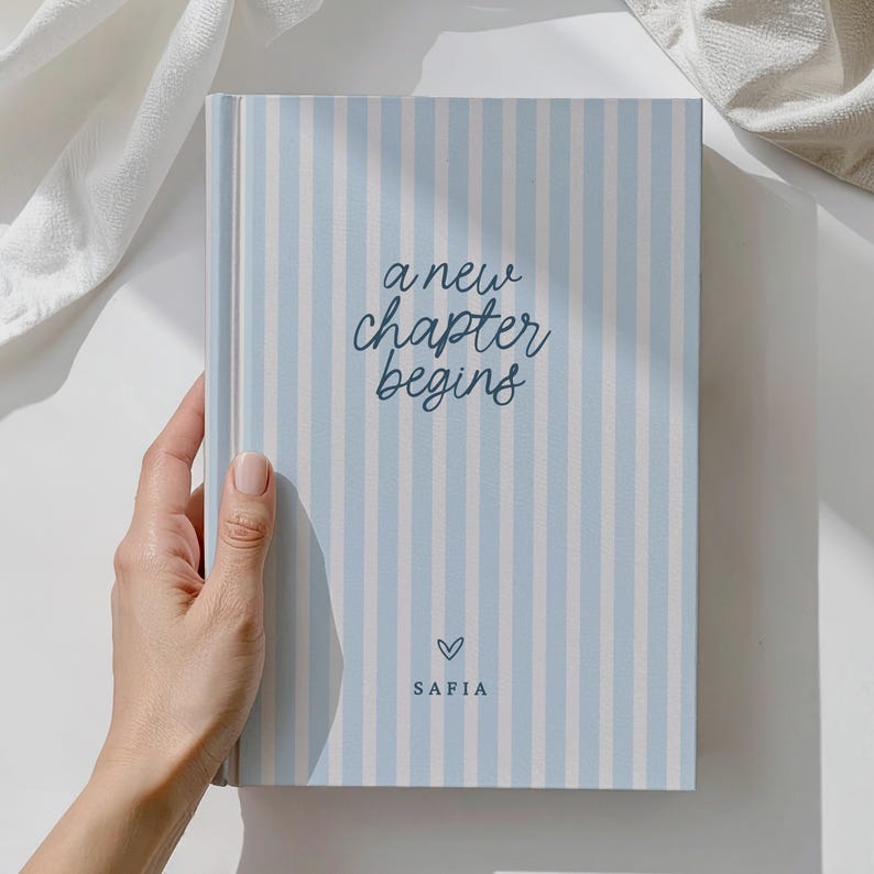 May include: A hardback journal with a light blue and white striped cover. The text "a new chapter begins" is written in cursive script, with a heart symbol and the name "SAFIA" below. The journal is held by a hand.