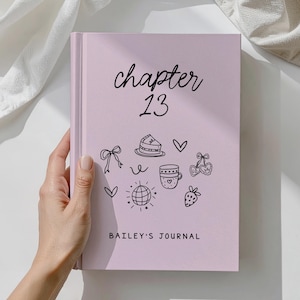 May include: A light pink journal with the text "chapter 13" in cursive and "BAILEY'S JOURNAL" in block letters. The cover features black line art of a bow, cake, hearts, a coffee cup, and a strawberry.