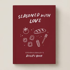May include: A deep red hardcover recipe book featuring the white text "SEASONED WITH LOVE" and "STEVEN & BAILEY'S RECIPE BOOK". The cover includes white line art illustrations of a tomato, a loaf of bread, pasta, and a fork.