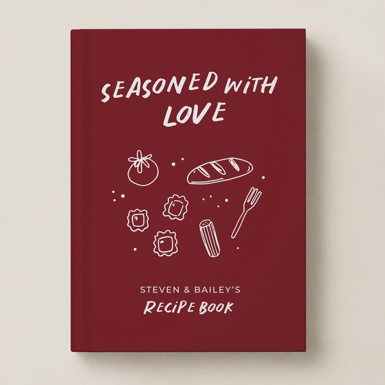 May include: A deep red hardcover recipe book featuring the white text "SEASONED WITH LOVE" and "STEVEN & BAILEY'S RECIPE BOOK". The cover includes white line art illustrations of a tomato, a loaf of bread, pasta, and a fork.