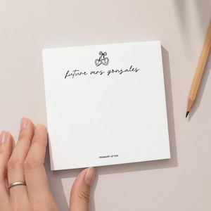 May include: A white square notepad with the text "future mrs gonzales" in a handwritten style, along with a small cherry illustration. The phrase "THOUGHT OF YOU" is printed at the bottom. A wooden pencil rests on the side.