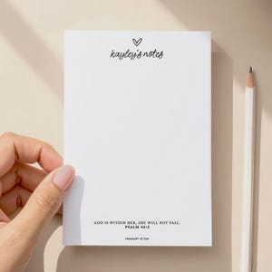 May include: A white notepad with the text "kayley's notes" and a heart symbol at the top. The bottom of the notepad has the text "GOD IS WITHIN HER, SHE WILL NOT FALL. PSALM 46:5 THOUGHT OF YOU". A pencil is next to the notepad.