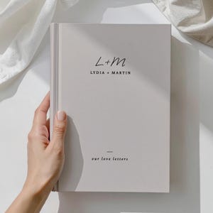 May include: A light gray book with the text "L+M LYDIA + MARTIN" in a handwritten font. Below, the text "our love letters" is printed in a smaller, sans-serif font. The book is held by a hand.