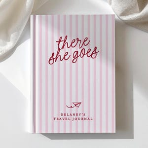 May include: A hardcover journal featuring a vertical light pink and white striped pattern. The front cover displays the dark red cursive text there she goes, a small red paper airplane icon, and the text DELANEY'S TRAVEL JOURNAL in red.