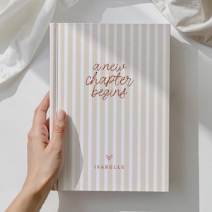 May include: A hardback journal with a beige and white striped design. The cover displays the text "a new chapter begins" in a brown script font, a small heart, and the name "Isabelle".