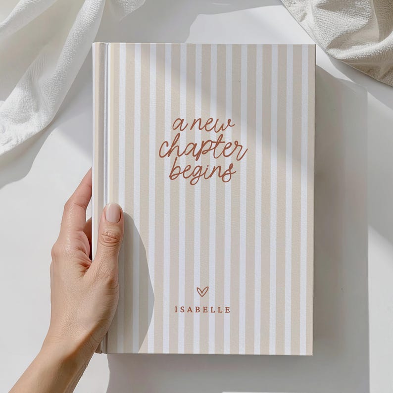 May include: A hardback journal with a beige and white striped design. The cover displays the text "a new chapter begins" in a brown script font, a small heart, and the name "Isabelle".