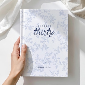 May include: A hardcover book with a floral pattern in light blue on a white background. The cover reads "CHAPTER thirty" in navy blue script, with the name "ANGELICA" below a heart symbol. The book is held by a hand.