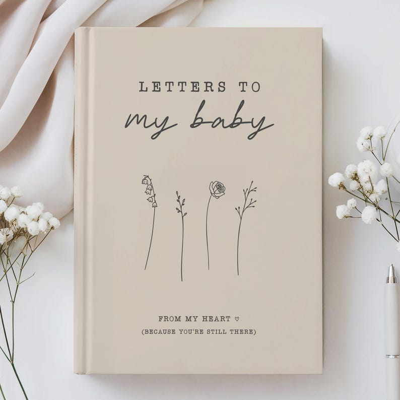 Op de afbeelding: Beigekleurig notitieboek met de tekst "Letters to my baby" en "From my heart (because you're still there)" in zwarte tekst. Het notitieboek heeft een eenvoudige lijntekening van vier bloemen.