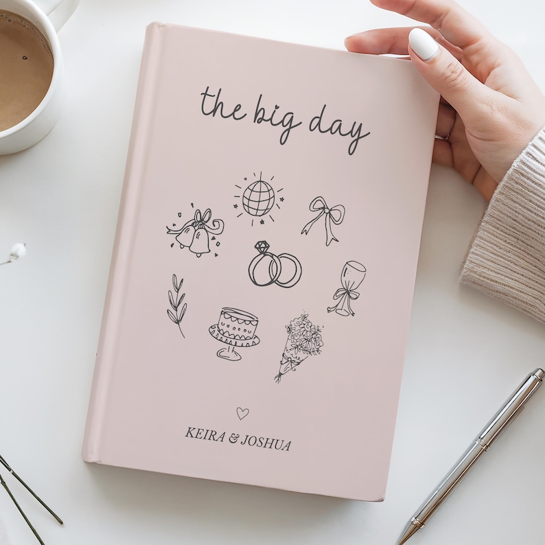 Pu&ograve; includere: Un diario di nozze rosa chiaro con le parole "the big day" in corsivo nero. La copertina presenta disegni a tratto nero di icone legate al matrimonio, tra cui campane, anelli e una torta. I nomi "Keira & Joshua" sono stampati in basso.