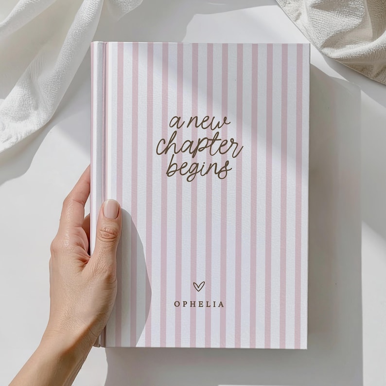 May include: A pink and white striped journal with the words "a new chapter begins" in brown script. A heart symbol and the name "Ophelia" are below the text. The journal is held by a hand, with a soft white cloth in the background.