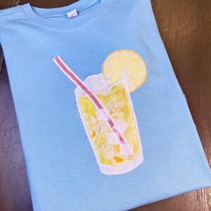 Puede incluir: Camiseta azul claro con un diseño bordado de una limonada, una rodaja de limón y una pajita a rayas rojas y blancas. La limonada es amarilla con cubitos de hielo. La camiseta está doblada sobre una superficie de madera.