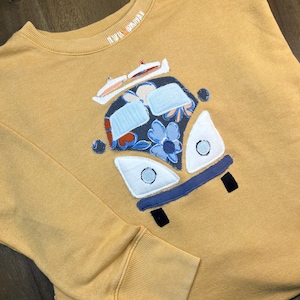 以下が含まれることがあります： マスタードイエローのトレーナーに、カラフルで模様のあるヴィンテージバンをモチーフにしたアップリケが施されています。バンは青と白の花柄で、白い屋根にはサーフボードが載っています。トレーナーの襟には「NEW TO ROAD」の文字が見えます。