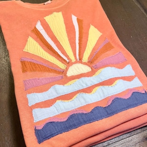 Puede incluir: Una camiseta naranja coral con un diseño de puesta de sol. El diseño está hecho de recortes de tela en tonos de rosa, naranja, amarillo y azul. El sol es un círculo amarillo con un centro blanco. Las olas son azules y blancas.