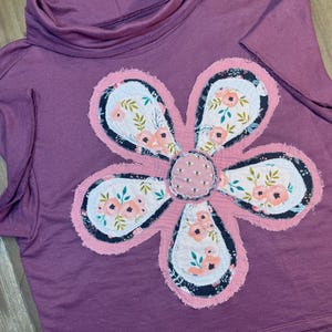 May include: A mauve colored shirt with a large pink and white floral applique. The flower is made of fabric scraps and has a patchwork design.