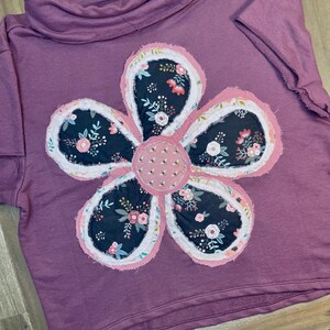 May include: A mauve colored long-sleeved shirt with a large, multi-colored flower applique on the front. The flower is made of fabric scraps in shades of pink, white, and black with a floral print.