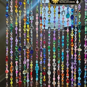 May include: A beaded curtain featuring multiple vertical strands of multicolored glass and acrylic beads in various shapes and sizes. A black rectangular logo with a gold star and the text Luxe SUNCATCHERS is visible near the top right.