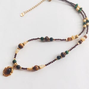 May include: A layered necklace featuring a pendant with a brown stone and gold-colored frame. The necklace has multiple strands of small, dark red beads, interspersed with brown, green, and cream-colored beads. The clasp is gold-colored.