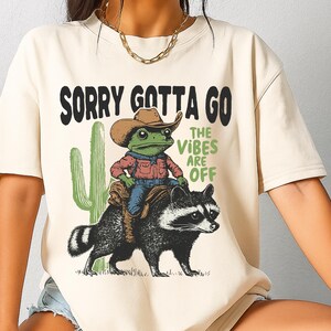 Może przedstawiać: Kremowy t-shirt z grafiką przedstawiającą zieloną żabę w brązowym kowbojskim kapeluszu i czerwonej koszuli, jadącą na czarno-białym szopie. Obok zielonego kaktusa znajduje się napis SORRY GOTTA GO oraz THE VIBES ARE OFF w kolorze czarnym i zielonym.