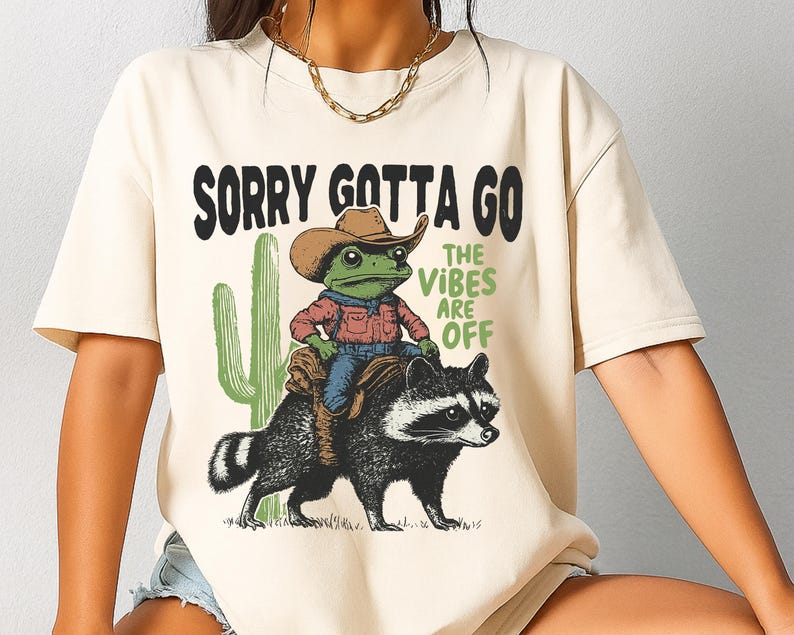 Może przedstawiać: Kremowy t-shirt z grafiką przedstawiającą zieloną żabę w brązowym kowbojskim kapeluszu i czerwonej koszuli, jadącą na czarno-białym szopie. Obok zielonego kaktusa znajduje się napis SORRY GOTTA GO oraz THE VIBES ARE OFF w kolorze czarnym i zielonym.