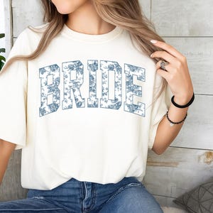 May include: A cream-colored short-sleeve t-shirt featuring the word BRIDE in large, block-style letters across the chest. The letters are filled with a blue and white floral pattern. The shirt has a crew neckline and a relaxed, loose fit.