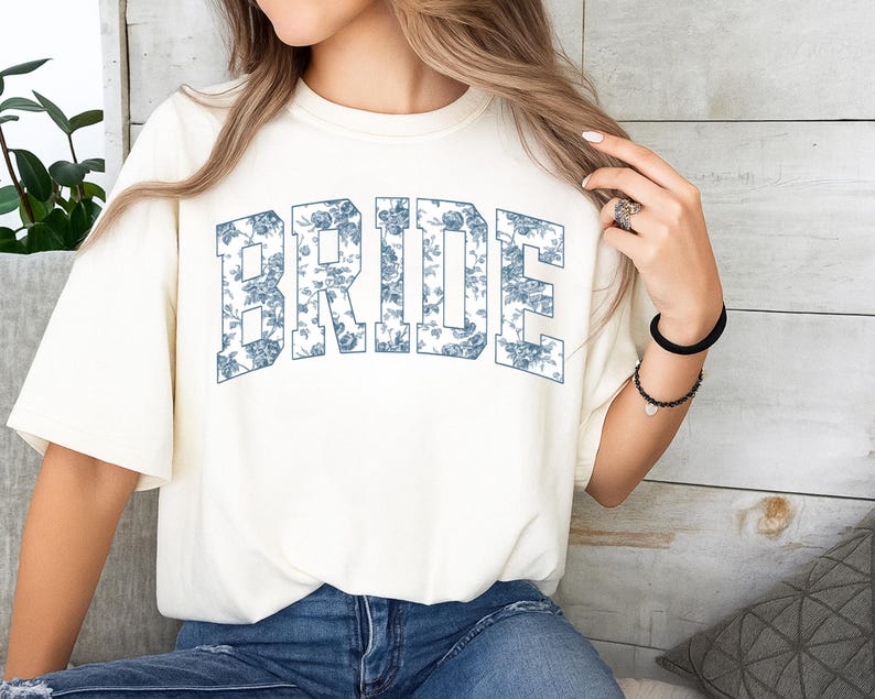 May include: A cream-colored short-sleeve t-shirt featuring the word BRIDE in large, block-style letters across the chest. The letters are filled with a blue and white floral pattern. The shirt has a crew neckline and a relaxed, loose fit.