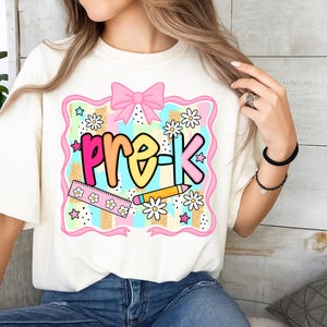 May include: Cream-colored t-shirt with a colorful "pre-K" graphic. The design includes a pink bow, stars, daisies, a ruler, and a pencil. The text is in a rainbow gradient, perfect for back-to-school.