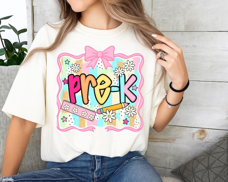 May include: Cream-colored t-shirt with a colorful "pre-K" graphic. The design includes a pink bow, stars, daisies, a ruler, and a pencil. The text is in a rainbow gradient, perfect for back-to-school.